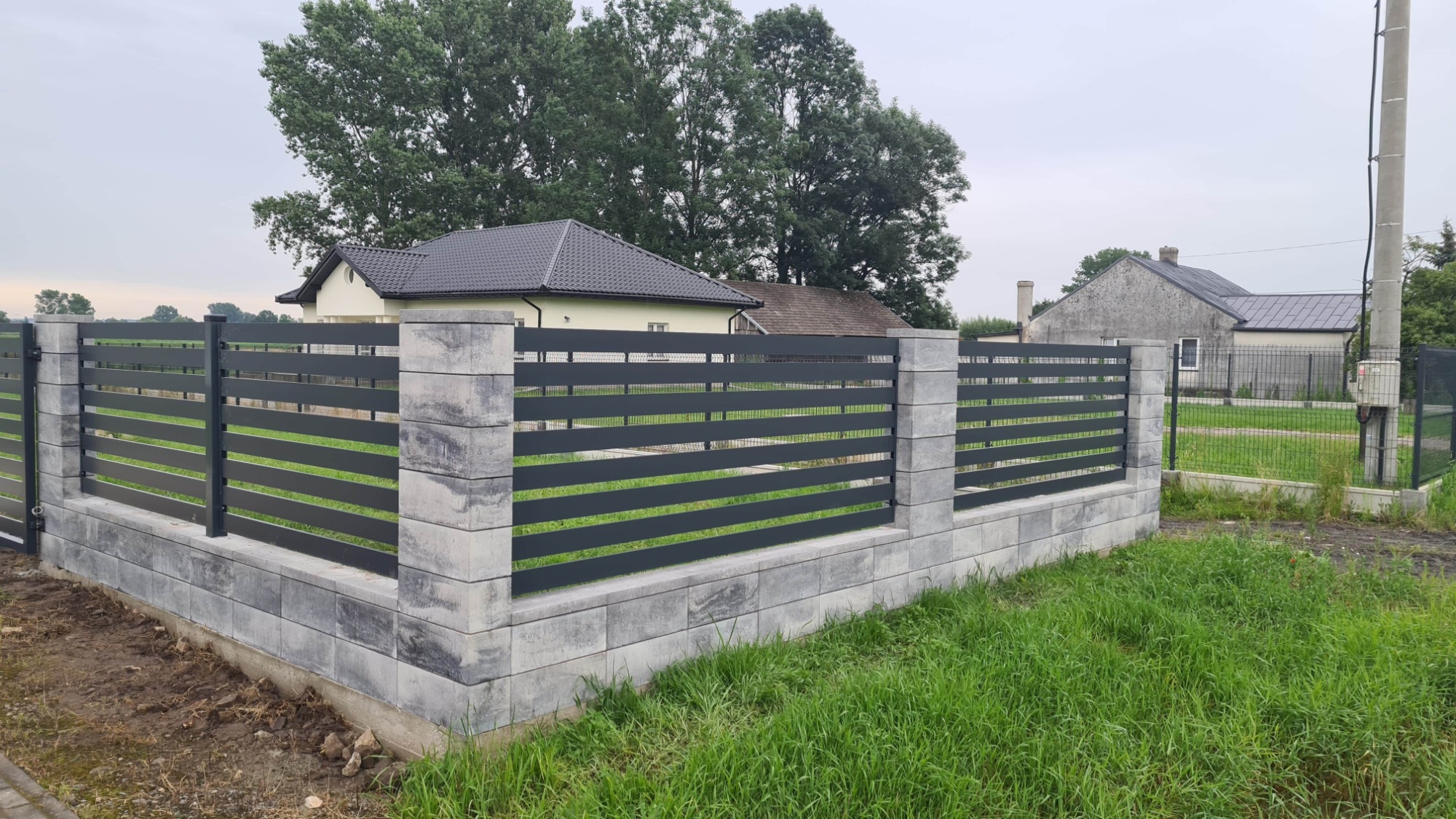 Nowoczesne ogrodzenie posesji z poziomych, grafitowych paneli zamontowanych na betonowych słupkach i podmurówce z szarej kostki, na tle zielonej trawy i zabudowań.