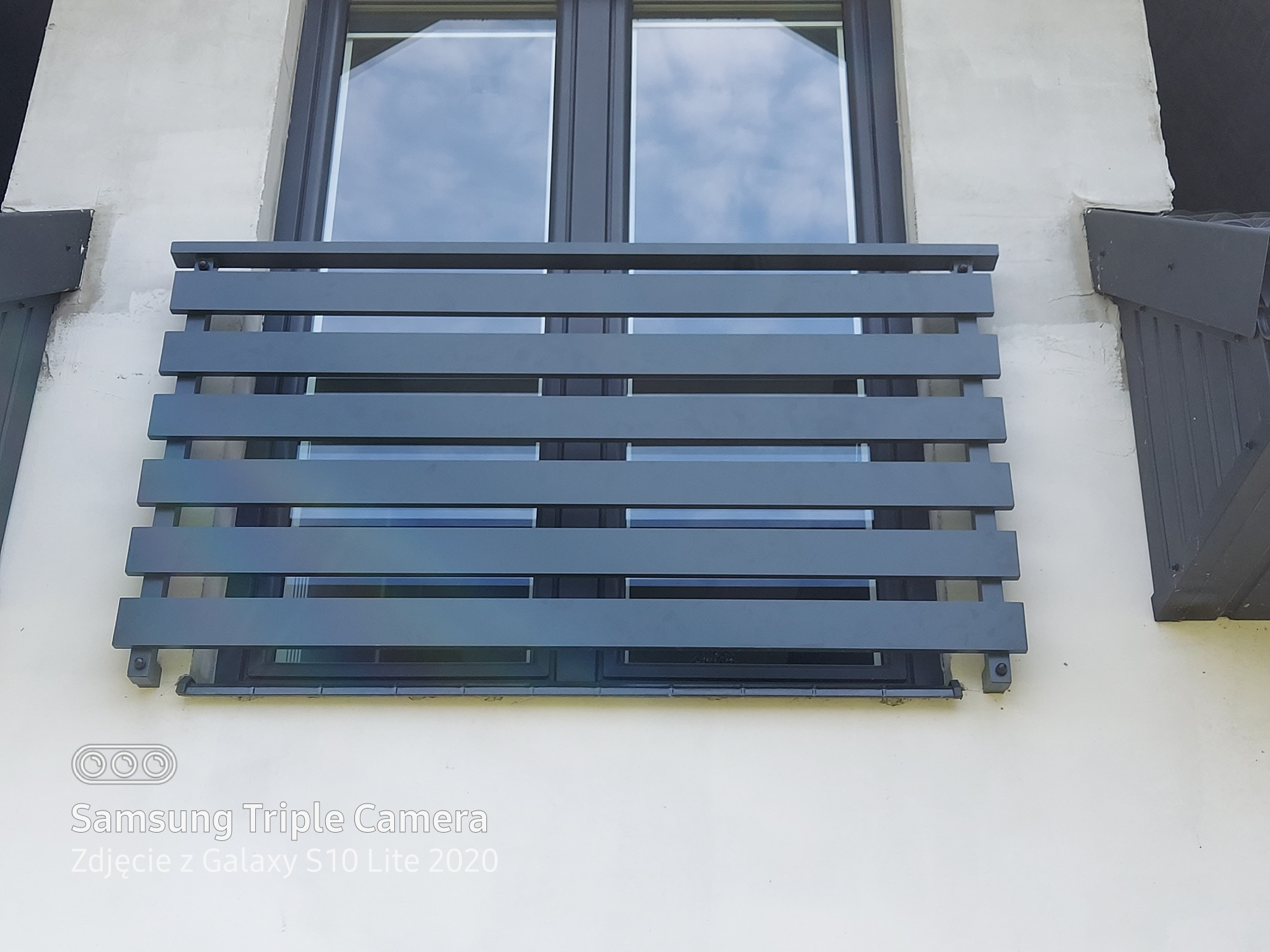 Szara, pozioma osłona okienna z metalowych listew zamontowana na elewacji budynku, widoczna na tle okna z odbijającym się niebem.