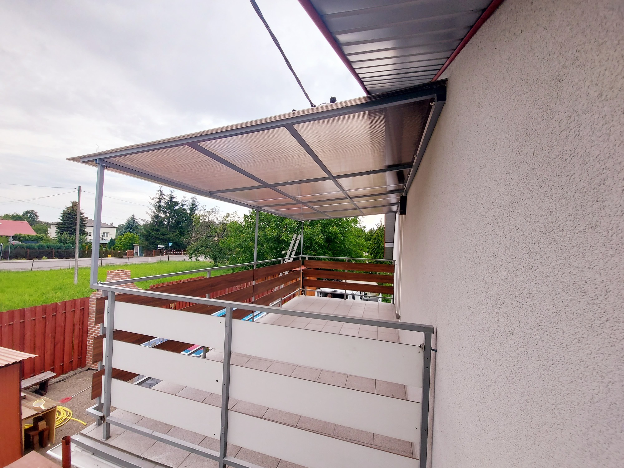 Zadaszony balkon z drewniano-białą balustradą, widok z boku, na zewnątrz szara pogoda.