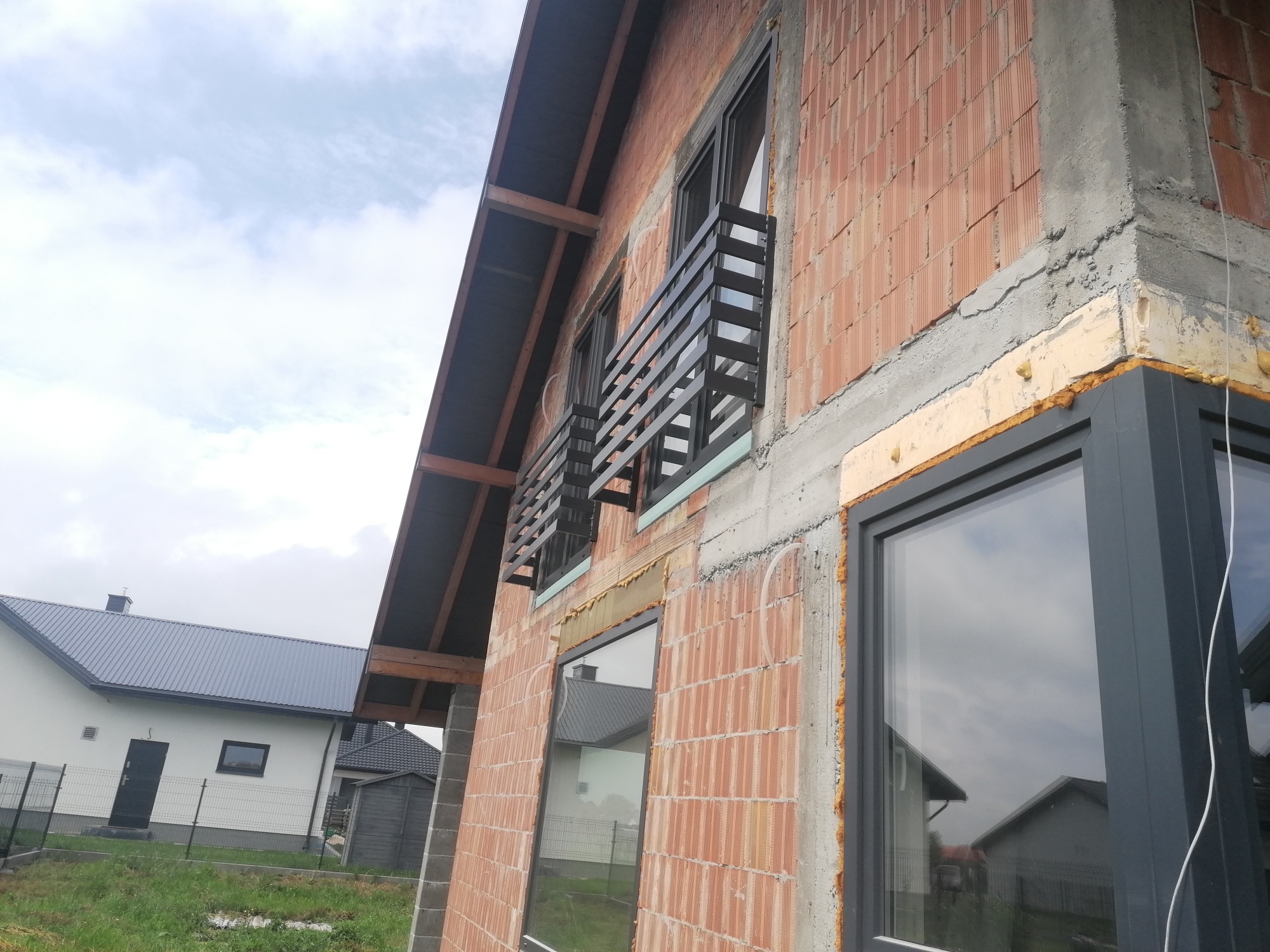 Surowy stan budynku w trakcie budowy z zamontowanymi czarnymi, poziomymi balustradami balkonowymi i szarymi oknami, widok z dołu na tle zachmurzonego nieba.