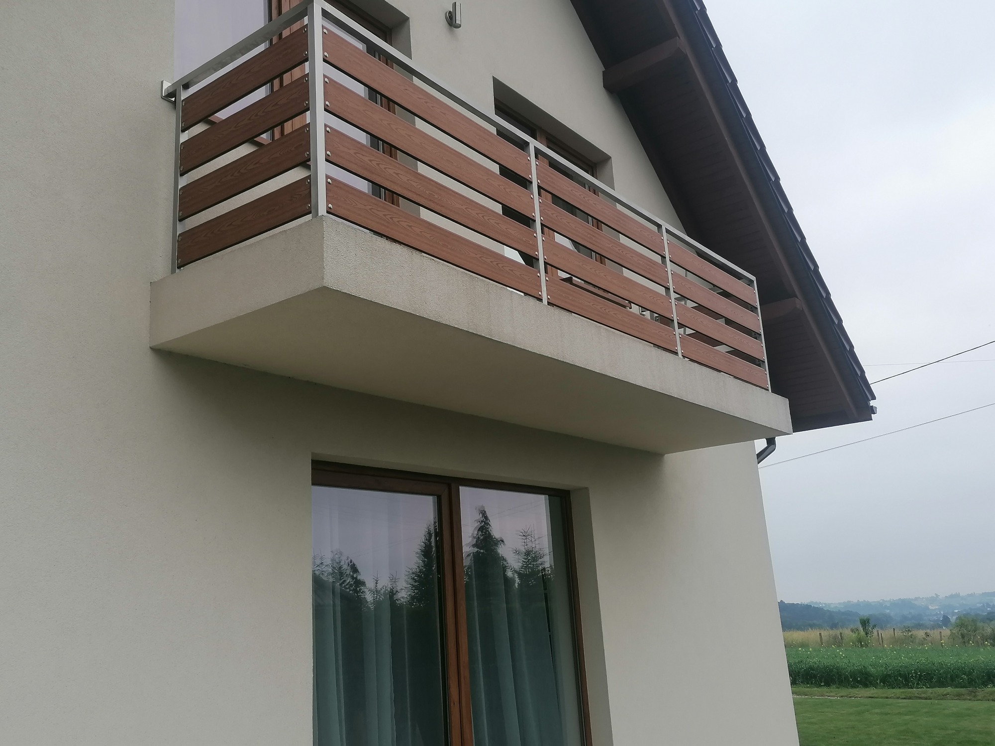 Balkon z nowoczesną balustradą z poziomych paneli imitujących drewno, osadzonych w metalowej ramie, widok z dołu na tle domu i krajobrazu.