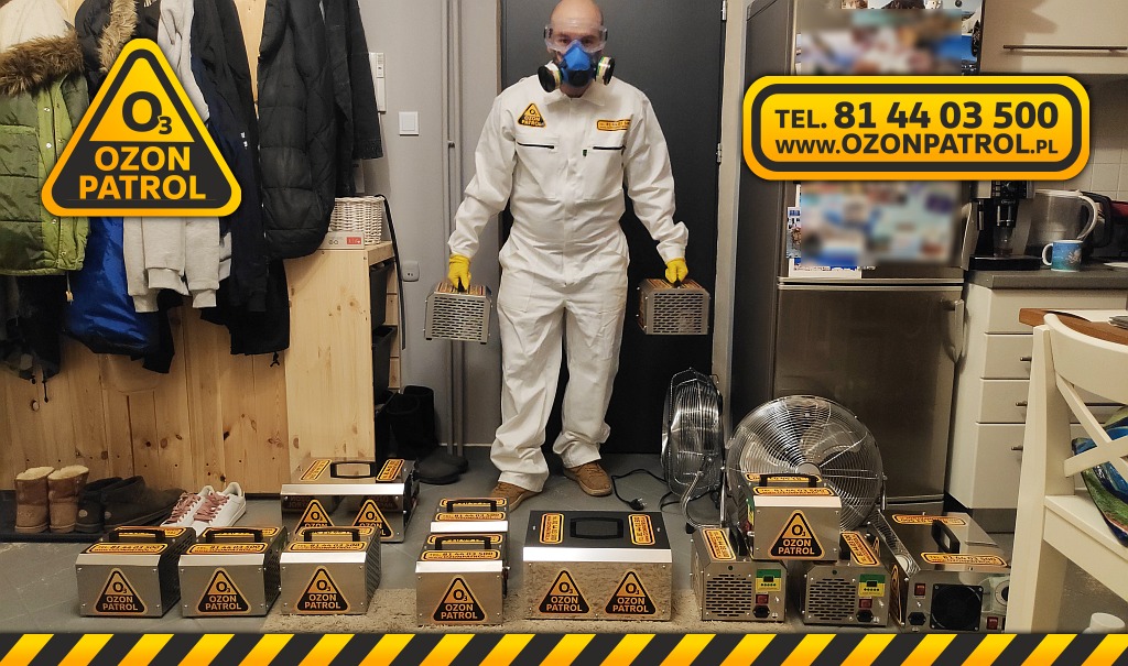 Pracownik w kombinezonie ochronnym i masce trzyma dwa ozonatory na tle pomieszczenia z ustawionymi na podłodze kolejnymi urządzeniami do ozonowania.