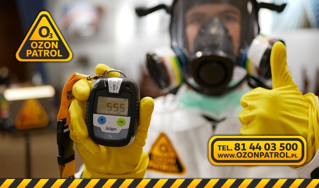 Pracownik w kombinezonie ochronnym i masce gazowej trzyma miernik stężenia ozonu Dräger, wyświetlający wartość 5.55 ppm, kciuk w żółtej rękawicy uniesiony w górę.
