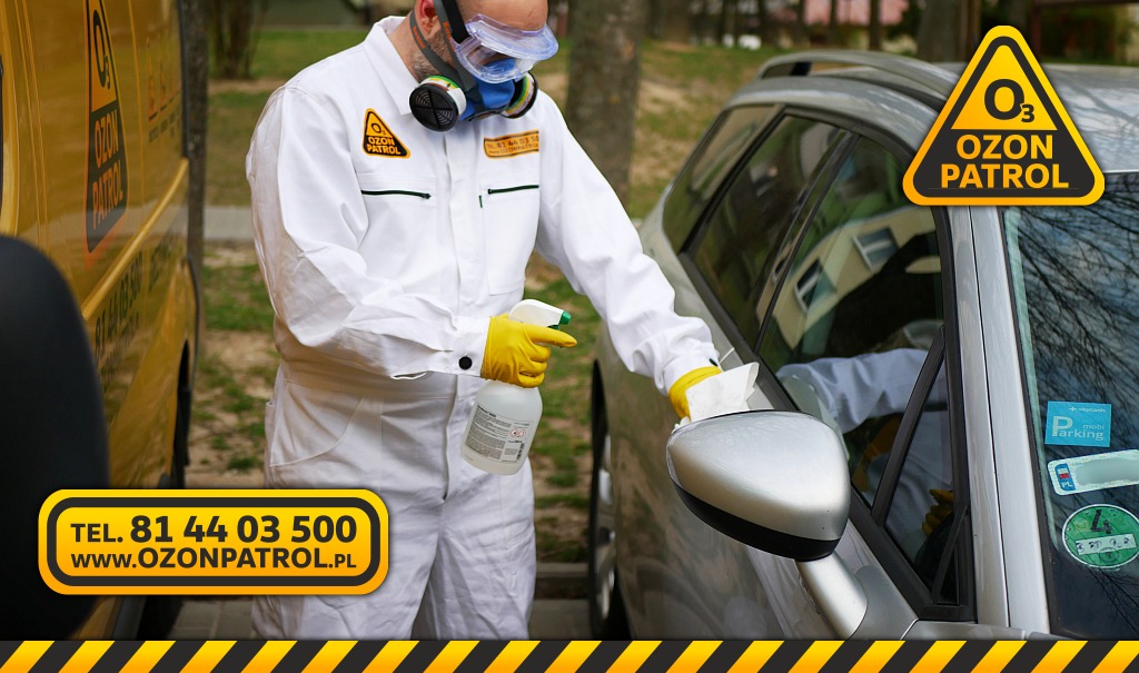 Pracownik w kombinezonie ochronnym, okularach i masce dezynfekuje lusterko srebrnego samochodu płynem z rozpylacza i białą szmatką. W tle żółty samochód z logo firmy.