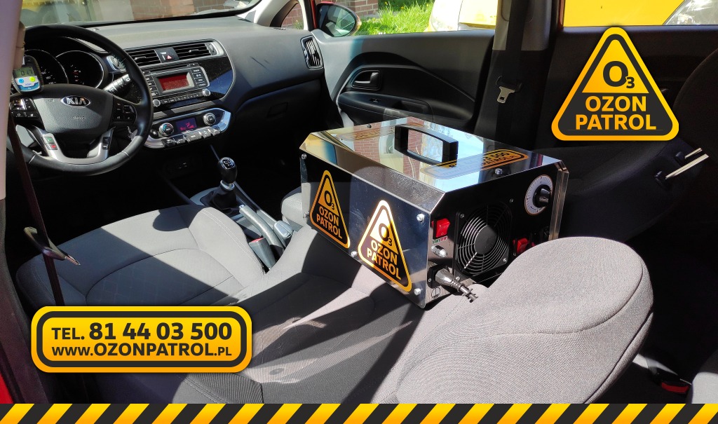 Ozonator przemysłowy umieszczony na tylnym siedzeniu samochodu osobowego marki Kia, podczas procesu odkażania wnętrza pojazdu. Na urządzeniu widoczne naklejki ostrzegawcze z logo firmy.