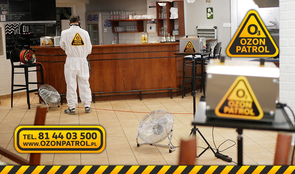 Pracownik w białym kombinezonie i masce ochronnej stoi w pomieszczeniu z drewnianym barem, obok wentylatory i urządzenia do ozonowania z naklejką 'Ozon Patrol', na ścianie menu i lodówka Pepsi.