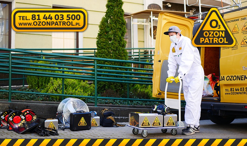 Pracownik w kombinezonie ochronnym i masce transportuje na wózku urządzenia do ozonowania, obok zaparkowany żółty samochód dostawczy z logo firmy Ozon Patrol i numerem telefonu, na chodniku leżą...