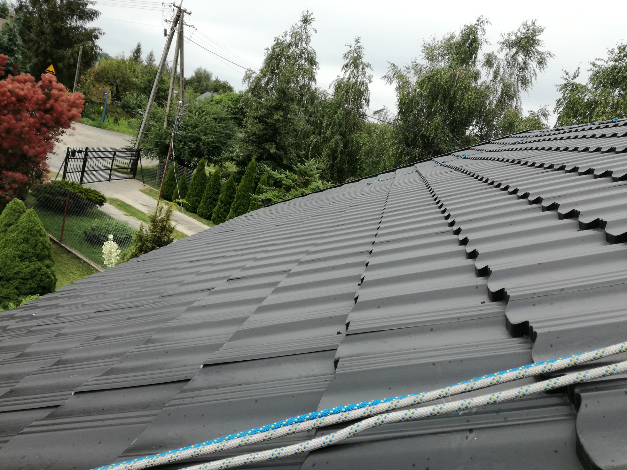 Nowy grafitowy dach z blachy trapezowej, widok z góry na tle zieleni i pochmurnego nieba, z fragmentem liny zabezpieczającej w prawym dolnym rogu.