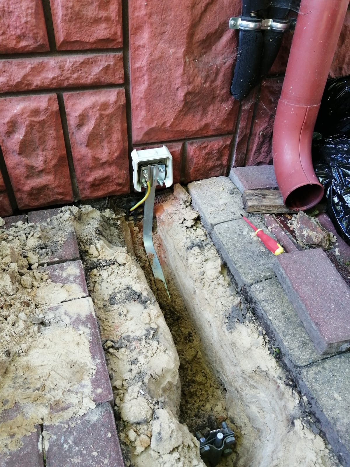 Przyłącze elektryczne do ściany budynku z uziemieniem, wykopany wąski rów wzdłuż fundamentu i kostki brukowej, widoczny przewód i śrubunek.
