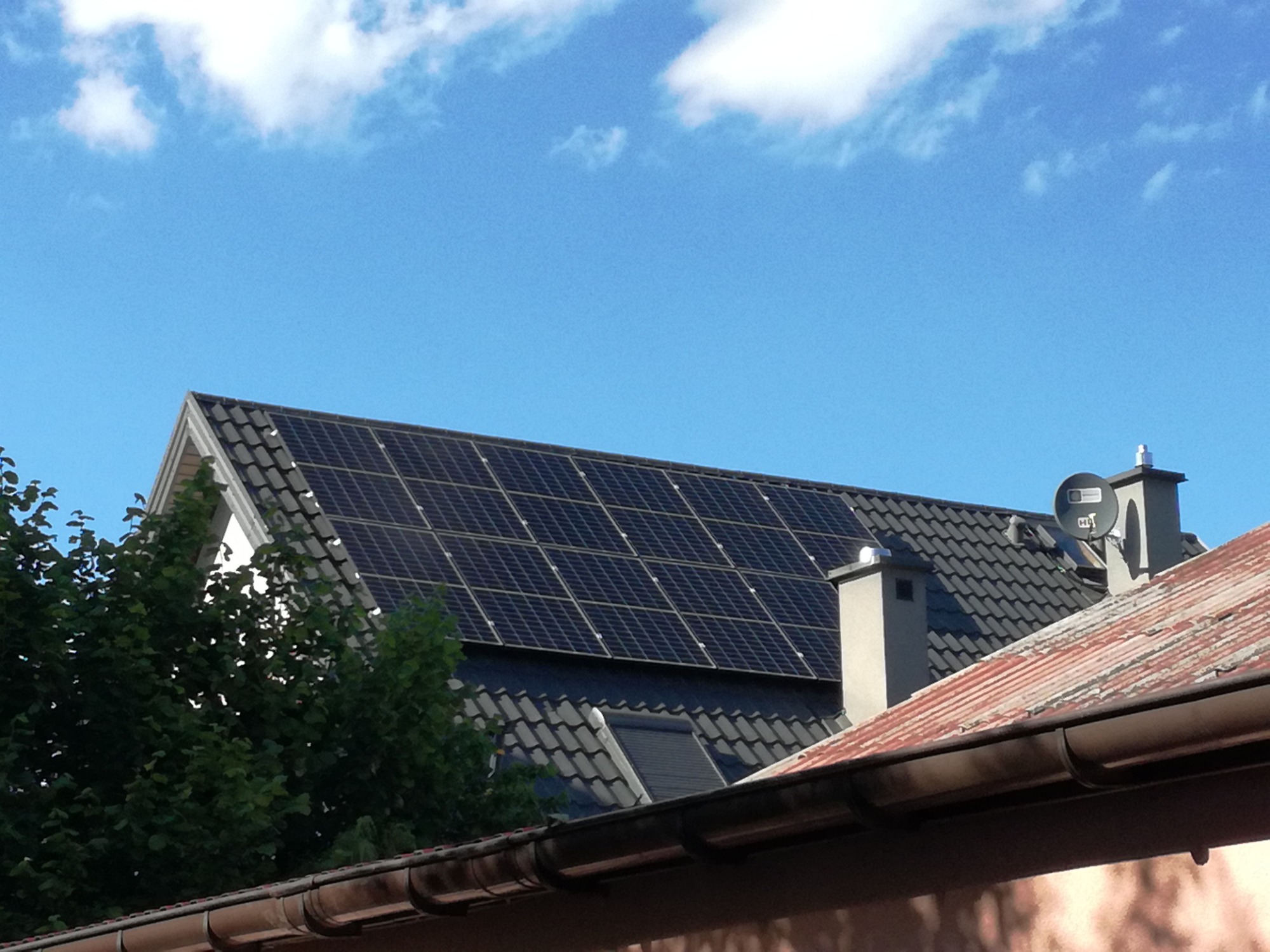 Panele słoneczne na ciemnym, spadzistym dachu z kominem, widoczne na tle błękitnego nieba z chmurami. Na pierwszym planie fragment dachu sąsiedniego budynku.