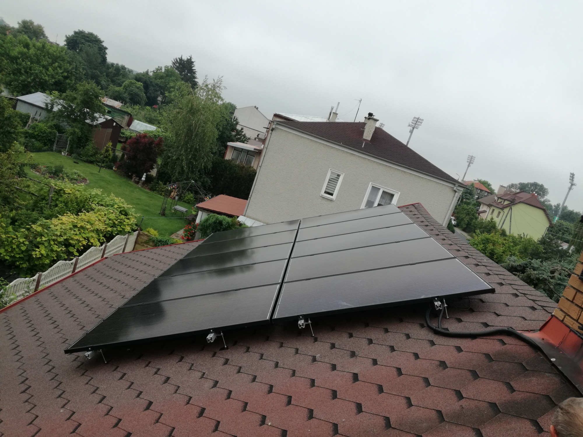 Cztery panele słoneczne zamontowane na dachu z brązowej dachówki bitumicznej, widok pod kątem, w tle domy i zieleń, pochmurne niebo.