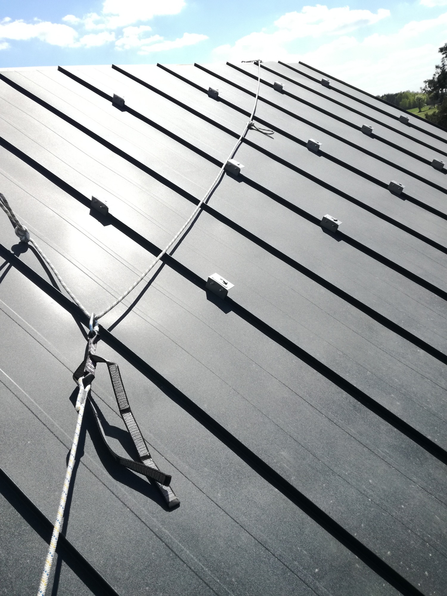 Szary dach z blachy trapezowej z zamontowanymi uchwytami do instalacji paneli słonecznych i liną zabezpieczającą z karabińczykiem.