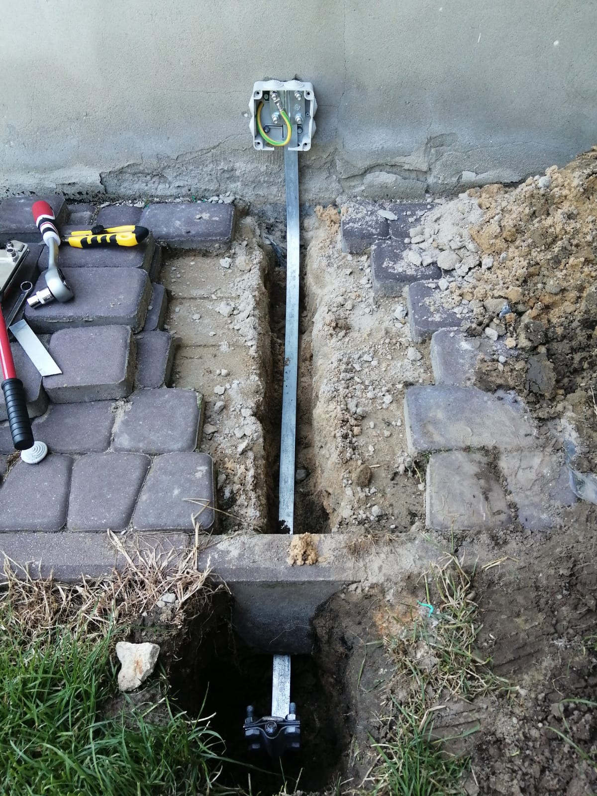 Montaż uziemienia instalacji: stalowa szyna uziemiająca w wykopie, skrzynka przyłączeniowa z przewodami ochronnymi, kostka brukowa i narzędzia obok.