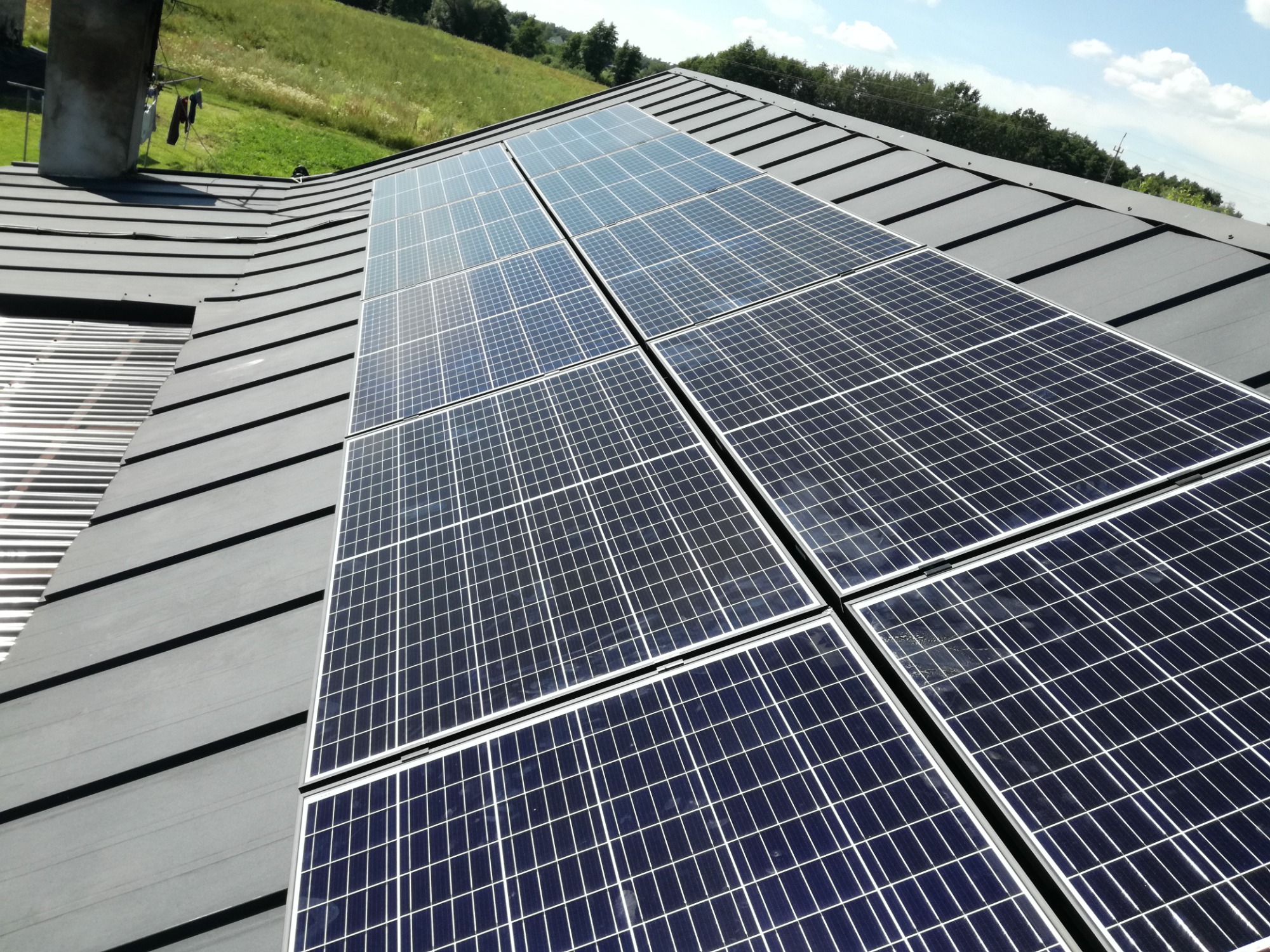 Panel fotowoltaiczny na grafitowym dachu blaszanym z widocznym kominem i zielonym wzgórzem w tle.