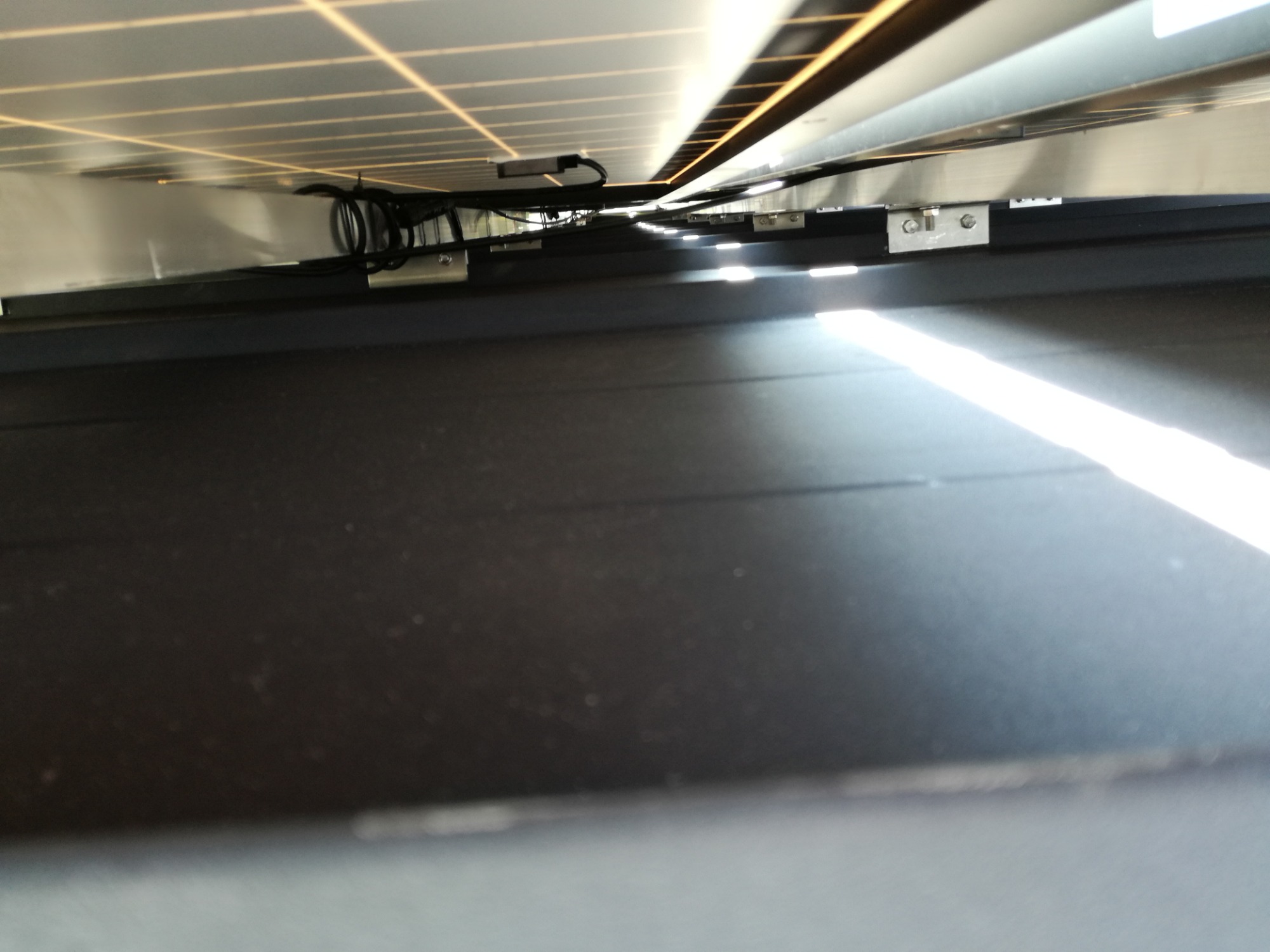 Widok od spodu na panele słoneczne z widocznymi kablami, mocowaniami i konstrukcją wsporczą, oświetlone promieniami słońca.