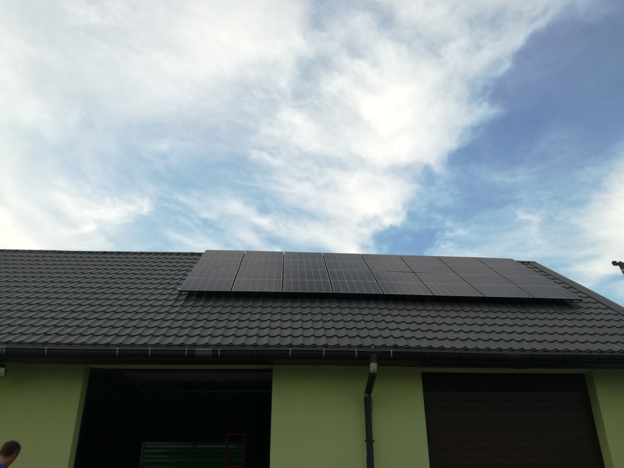 Panele fotowoltaiczne zamontowane na ciemnym, metalowym dachu budynku gospodarczego z jasnozieloną elewacją, widoczne na tle błękitnego nieba z białymi chmurami.