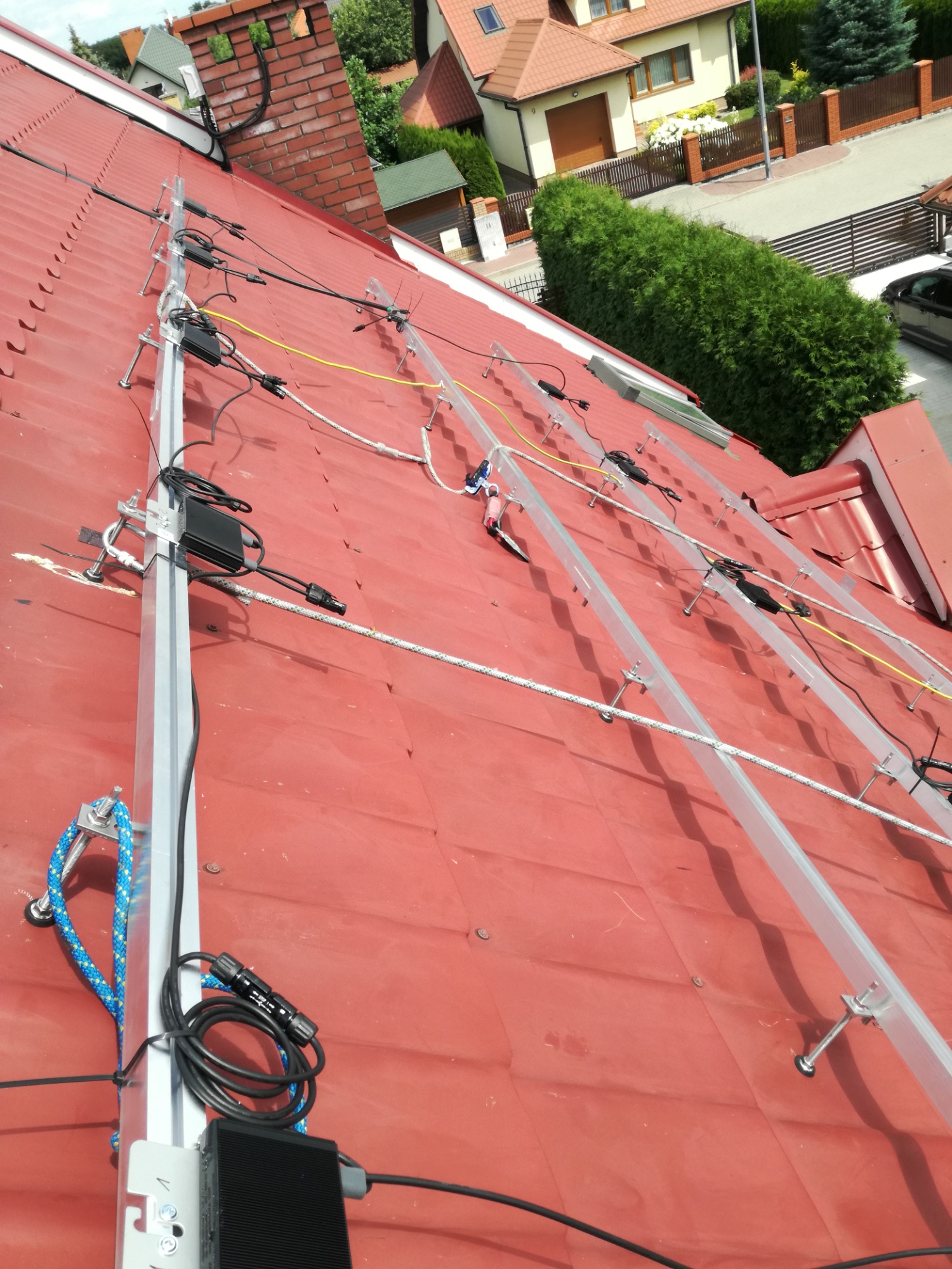 Montaż konstrukcji wsporczej pod panele słoneczne na czerwonym dachu z blachy trapezowej, widoczne kable i elementy mocujące.