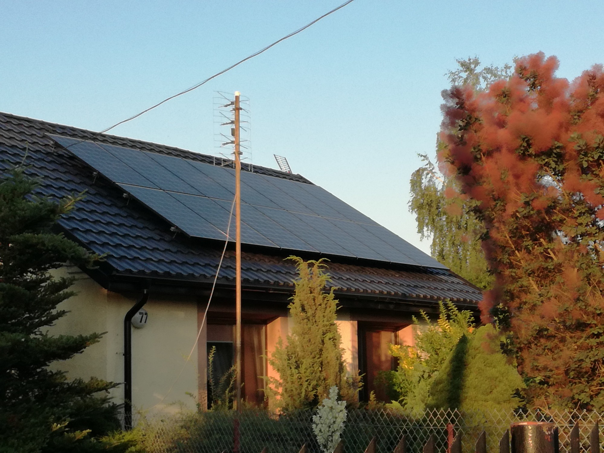 Panele słoneczne na dachu domu jednorodzinnego z anteną telewizyjną, widok z poziomu ogrodu z drzewami w tle.