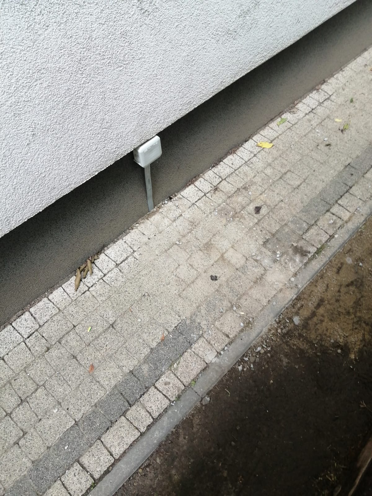Biała puszka elektryczna z szarą rurką przy ścianie budynku, widoczna na tle kostki brukowej.
