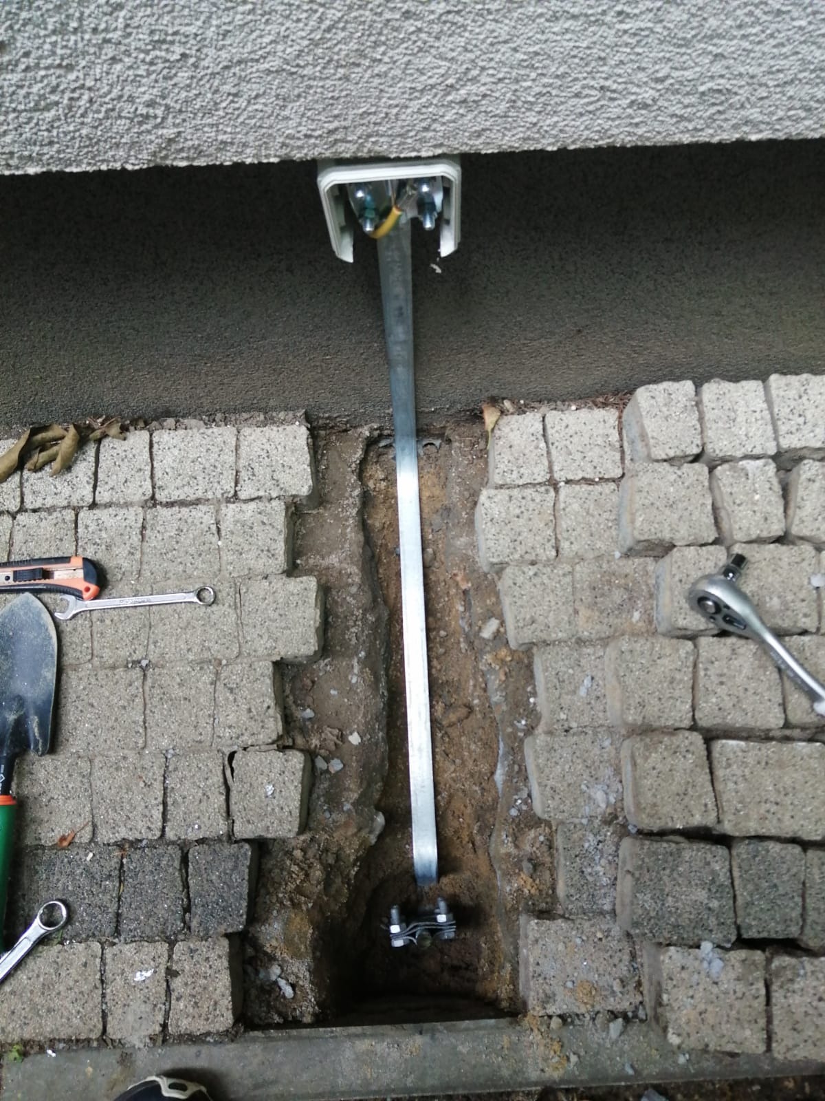 Montaż konstrukcji wsporczej, prawdopodobnie pod panele słoneczne, w Lublinie. Widoczny fragment ściany budynku, wykop pod fundament, narzędzia: klucze, łopata i nóż.