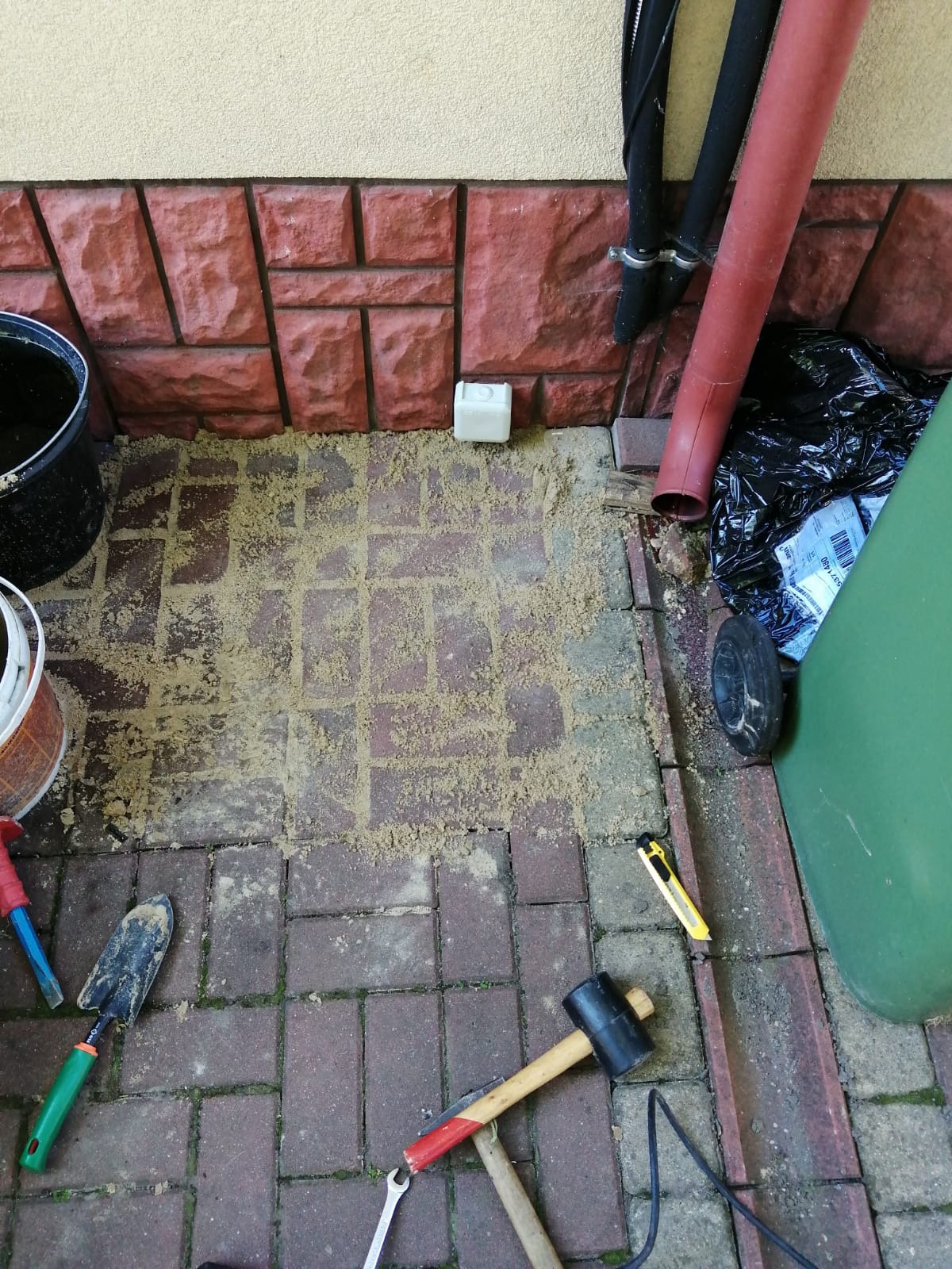 Fragment brukowanej powierzchni z piaskiem, narzędziami (szpachelka, młotek, klucz) i elementami instalacji przy ścianie budynku.