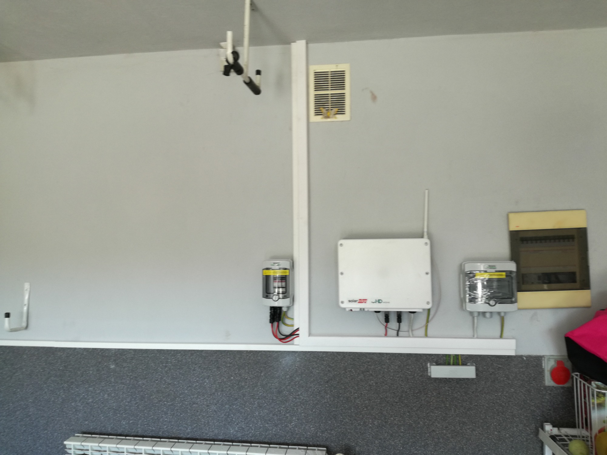 Szare pomieszczenie z zamontowanymi elementami instalacji elektrycznej, w tym biała skrzynka z anteną, zabezpieczenia nadprądowe i rozdzielnica elektryczna na ścianie.