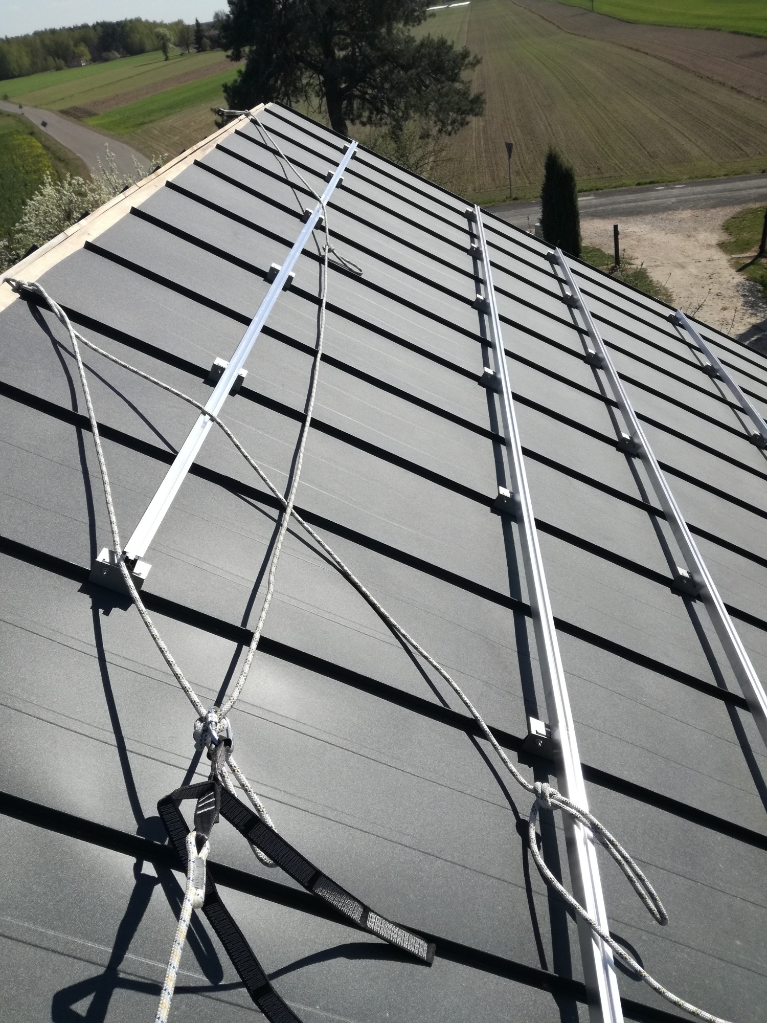 Montaż szyn pod panele słoneczne na dachu z użyciem zabezpieczeń linowych, widok z góry na dach i pola w tle.