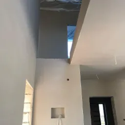 Wnętrze w trakcie prac wykończeniowych: gładkie, jasne ściany po nałożeniu tynku maszynowego, widoczne otwory na instalacje elektryczne i fragment izolacji stropu.