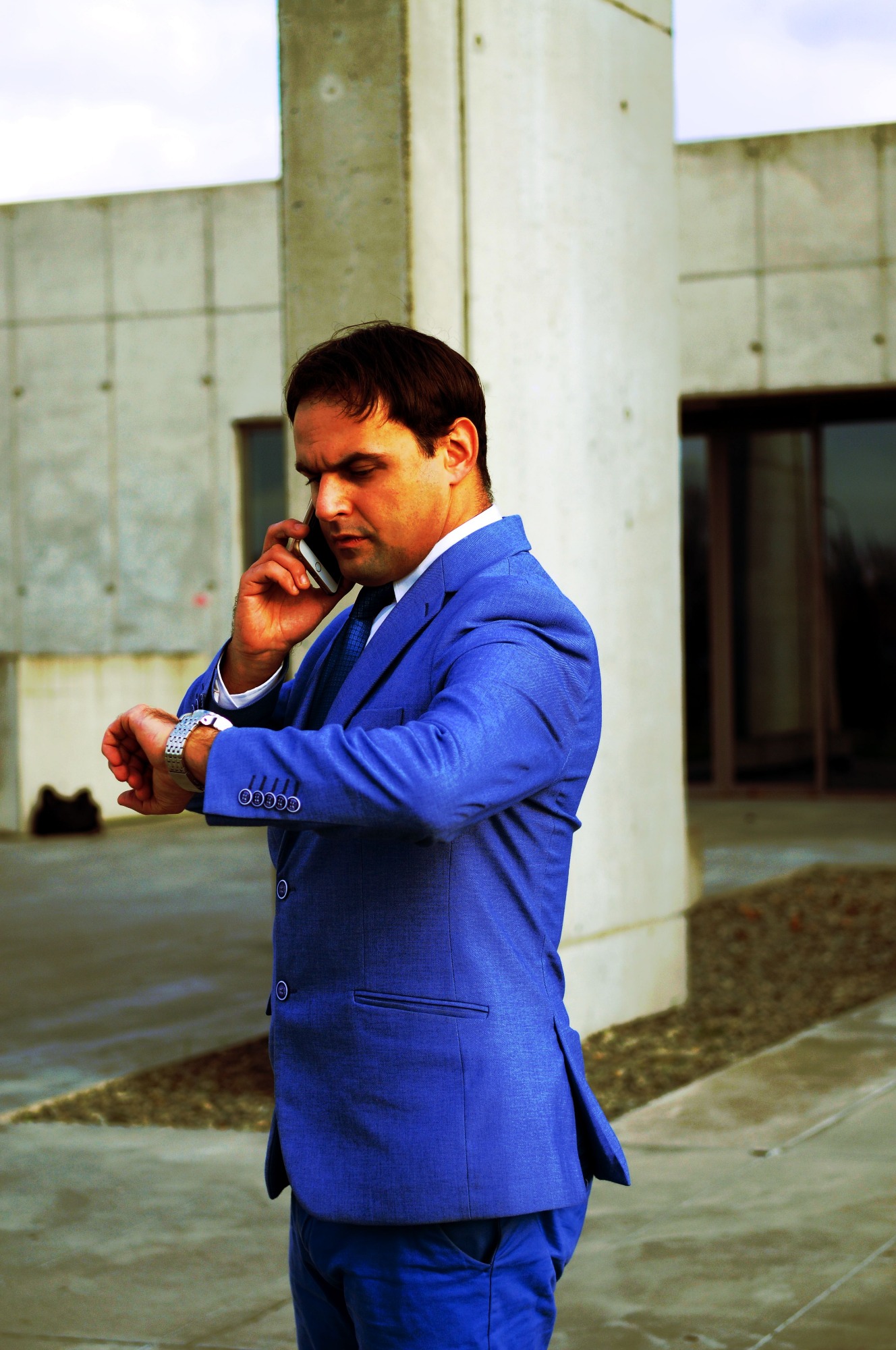 Mężczyzna w niebieskim garniturze rozmawia przez telefon, spoglądając na zegarek na tle nowoczesnego budynku.