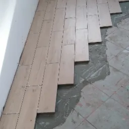 Układanie podłużnych, drewnopodobnych płytek z białymi krzyżykami dystansowymi na podłodze w trakcie remontu, widoczne stare płytki i zaprawa.