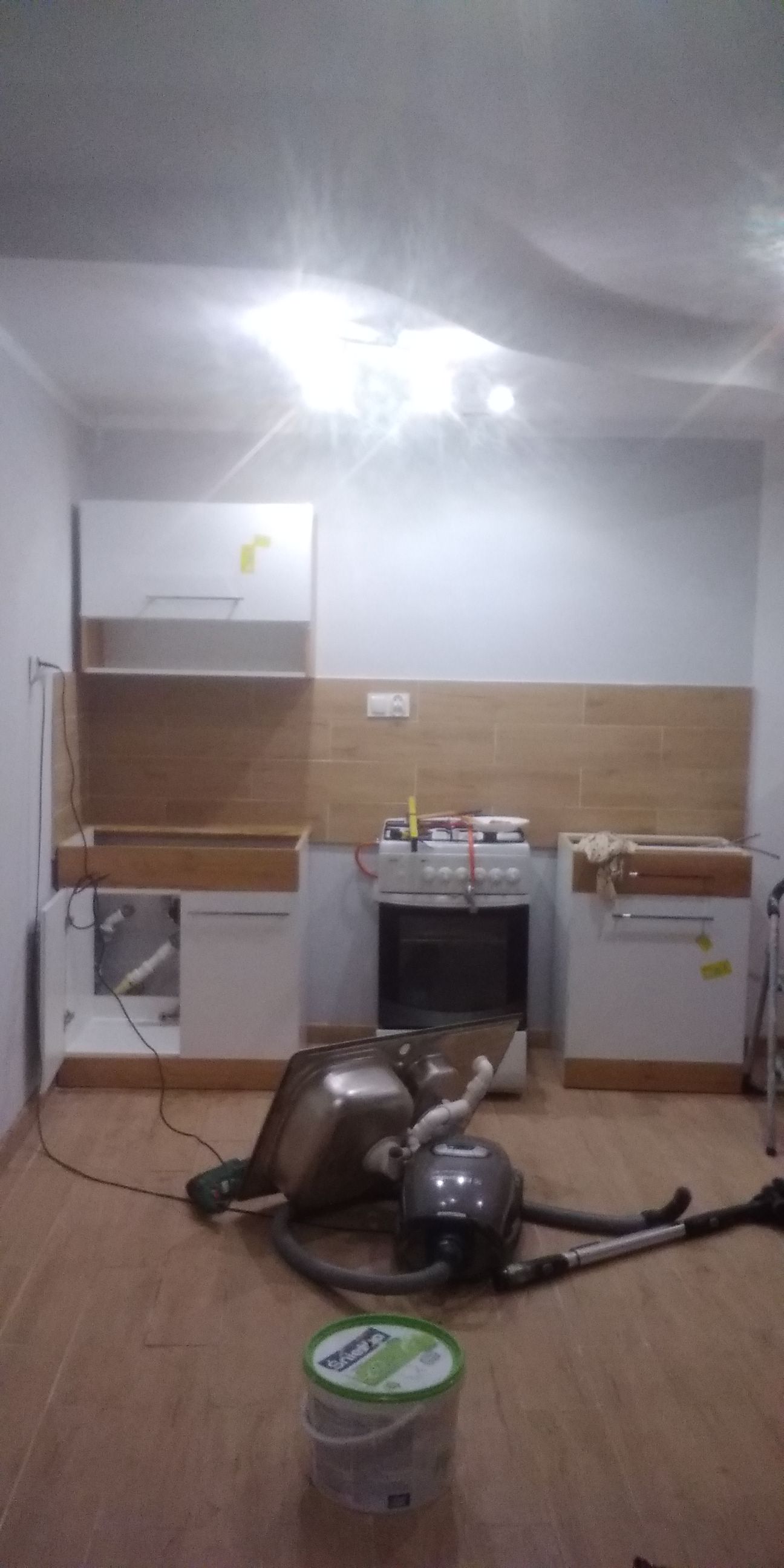 Wnętrze kuchni w trakcie montażu: widoczne niedokończone szafki z drewnianymi blatami, płytki na ścianie, zlew, odkurzacz i wiadro z farbą na podłodze.