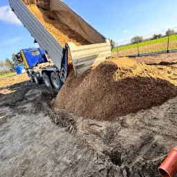 Ciężarówka wyładowuje piasek na placu budowy, widoczne ślady opon i rura kanalizacyjna.