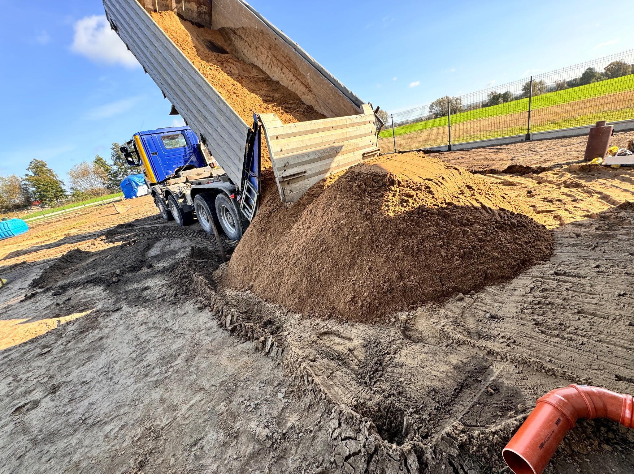 Ciężarówka wyładowuje piasek na placu budowy, widoczne ślady opon i rura kanalizacyjna.