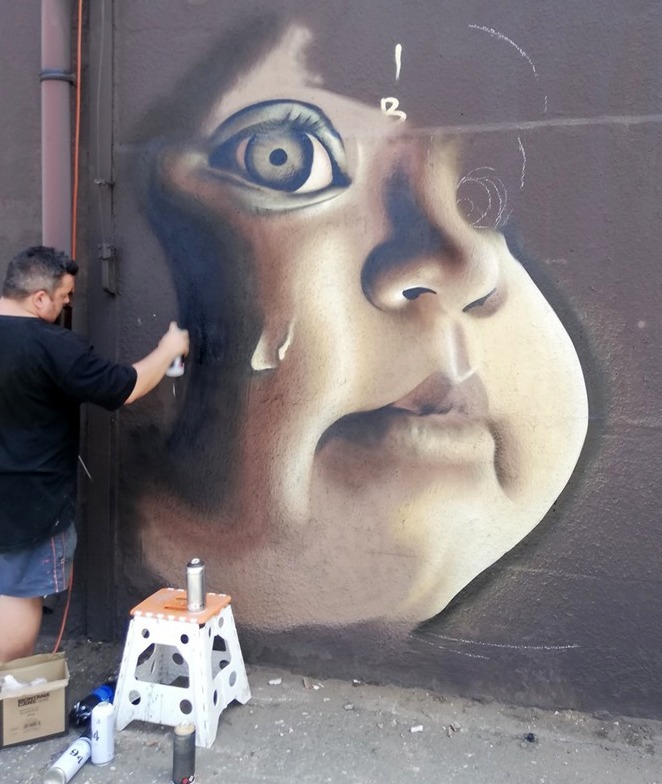 Artysta maluje portret dziecka z łzą na ścianie budynku za pomocą farby w sprayu, widoczny stołek z puszkami farby i karton na ziemi.