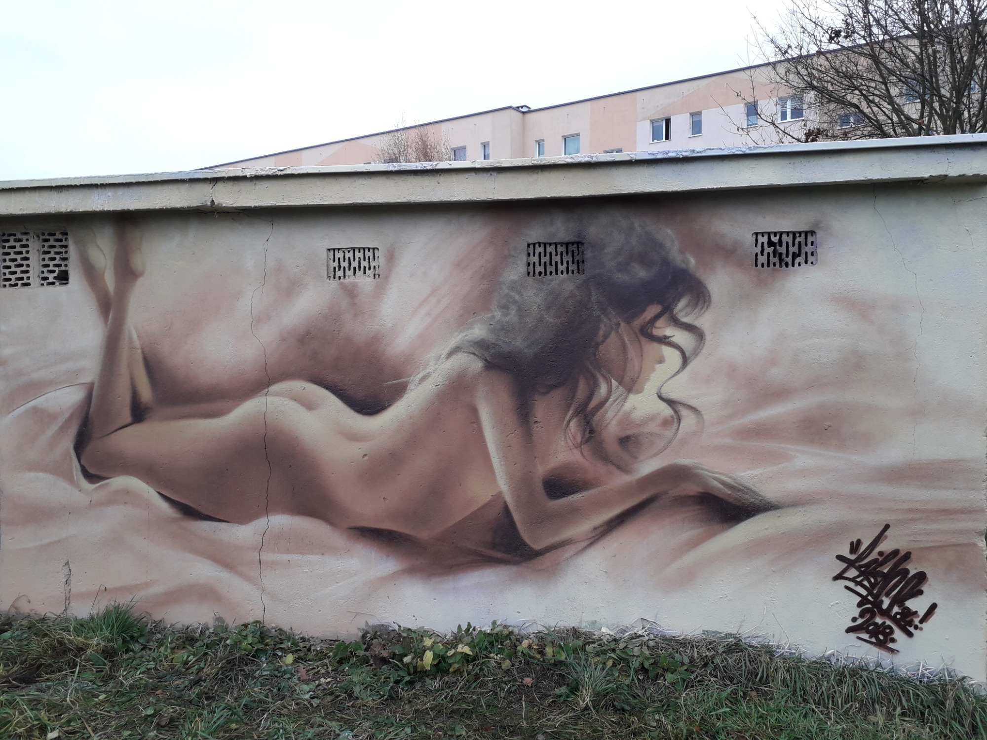 Graffiti przedstawiające nagą kobietę leżącą na boku, namalowane na betonowej ścianie z kratkami wentylacyjnymi, z podpisem artysty w prawym dolnym rogu.