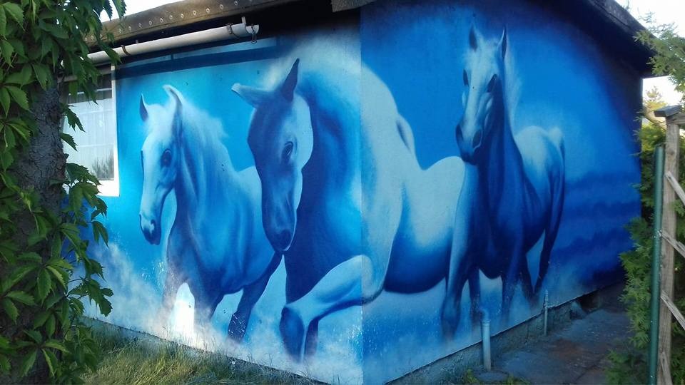 Ściana budynku ozdobiona błękitnym muralem przedstawiającym trzy galopujące konie.