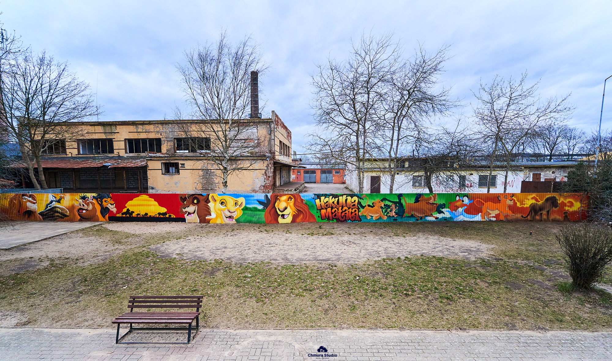 Kolorowy mural z motywami z Króla Lwa na ogrodzeniu w Lęborku, widok z perspektywy ławki, na tle budynków i drzew bez liści.