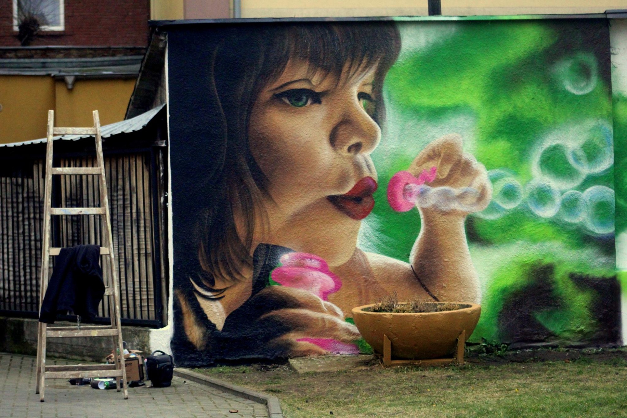 Mural przedstawiający portret kobiety dmuchającej bańki mydlane; widoczna drabina, puszki farb i torba malarska obok.