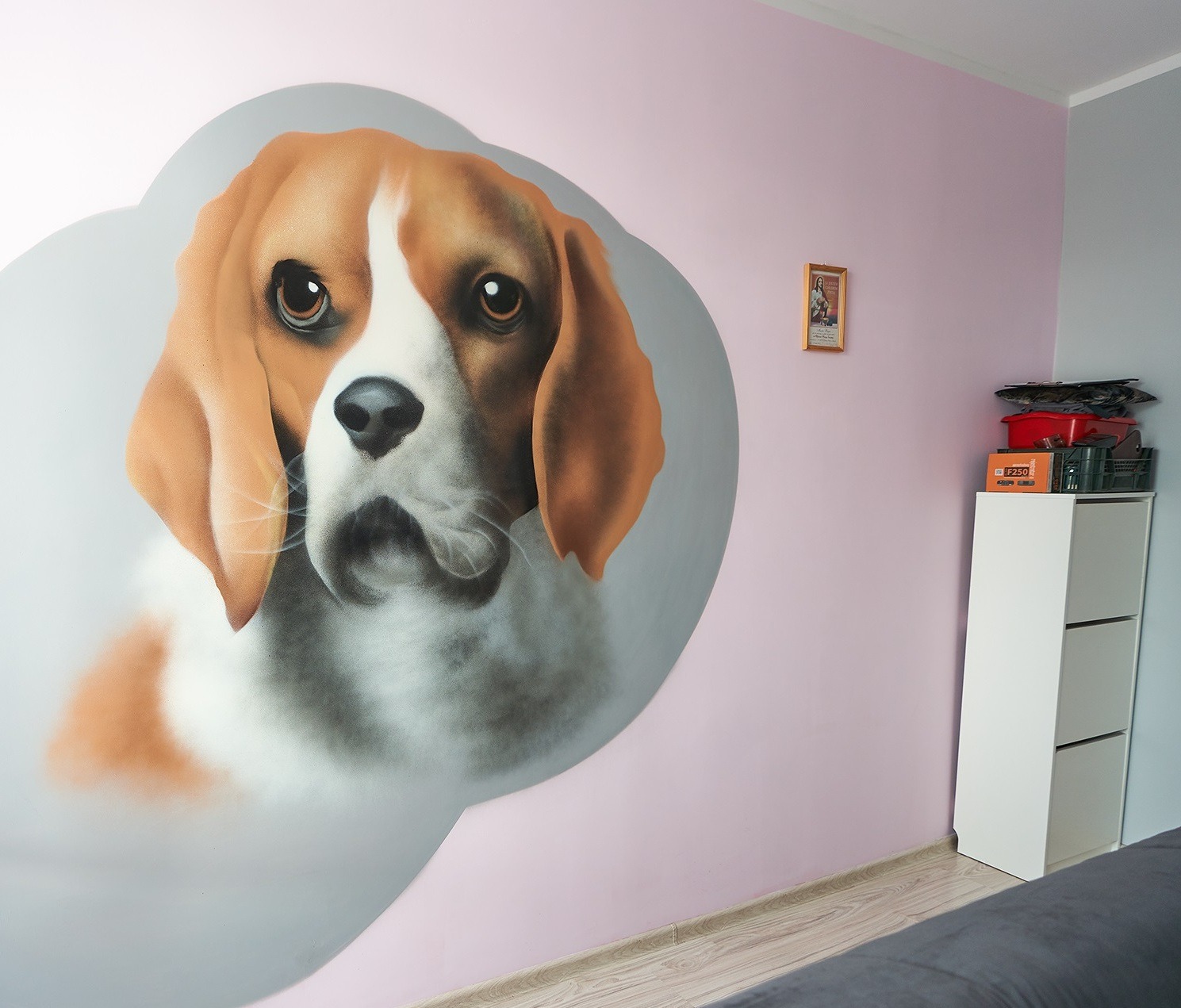 Portret psa rasy Beagle, namalowany bezpośrednio na ścianie w pokoju o pastelowych barwach, z widoczną szafką i obrazkiem religijnym w tle.