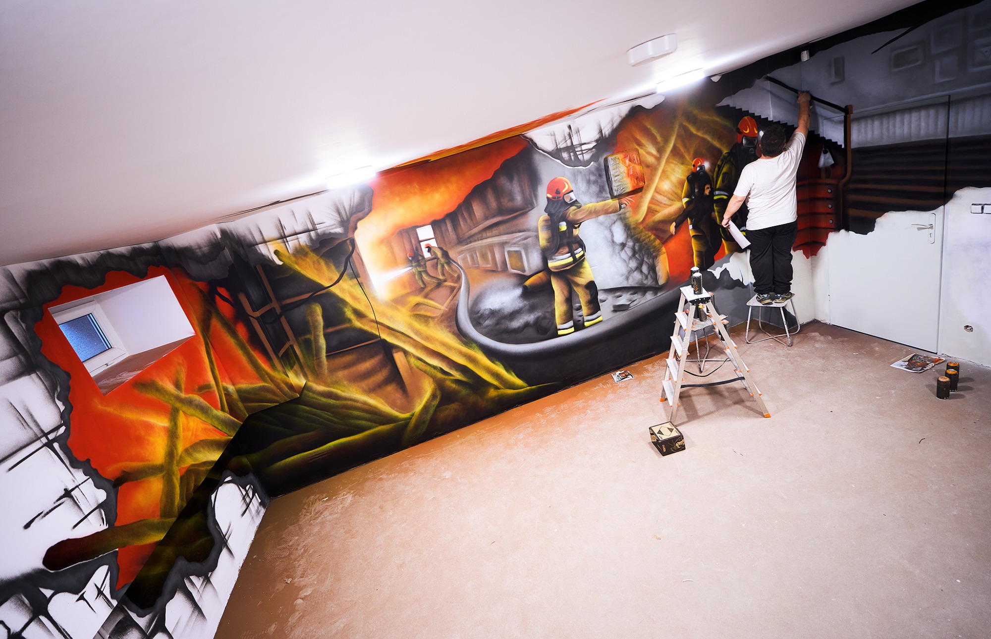Artysta na drabinie kończy malowidło ścienne przedstawiające strażaków gaszących pożar, widoczne puszki z farbą w sprayu.