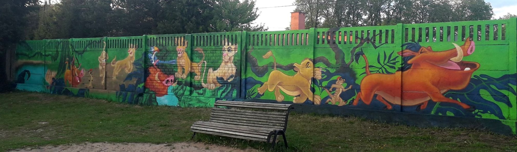 Barwny mural na betonowym ogrodzeniu przedstawiający postacie z filmu Król Lew, z ławką na pierwszym planie.