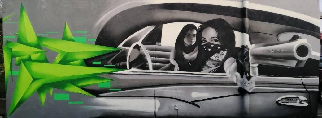 Graffiti przedstawiające dwie kobiety w samochodzie, jedna z pistoletem, z abstrakcyjnymi zielonymi kształtami po lewej stronie.