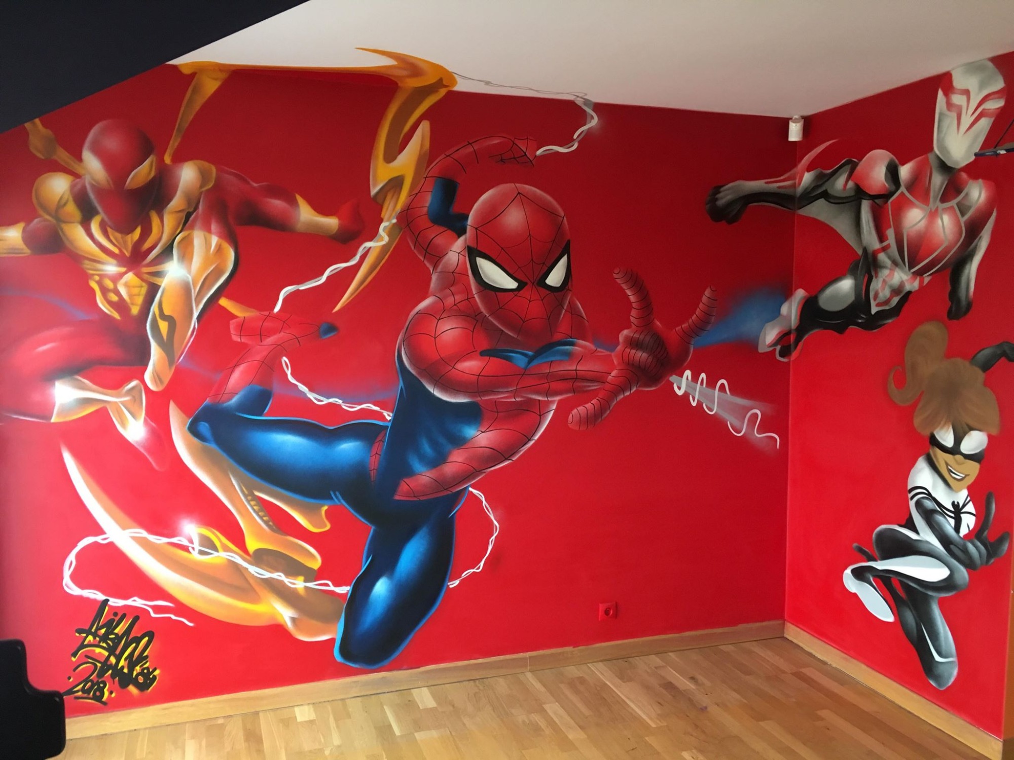 Pokój z czerwoną ścianą, na której namalowano postaci Spidermanów w różnych wersjach i stylach komiksowych, na podłodze parkiet.