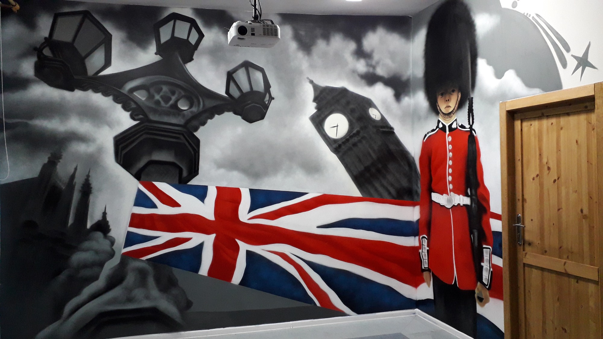 Ściana z ręcznie malowanym muralem w stylu brytyjskim: Big Ben, lampiony uliczne, flaga Wielkiej Brytanii i gwardzista w czerwonym mundurze, widoczny projektor pod sufitem.
