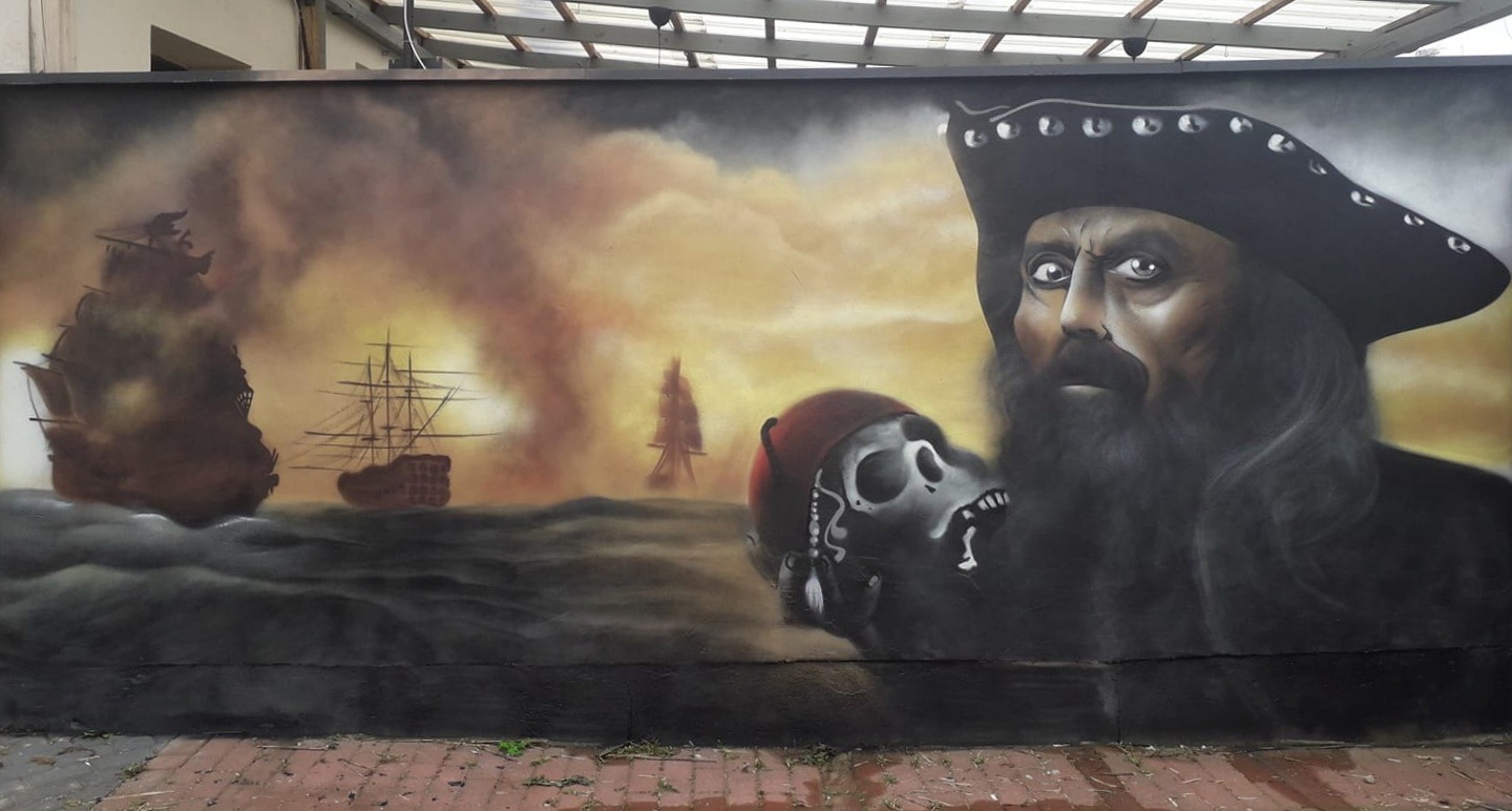 Mural przedstawiający pirata Czarnobrodego z czaszką w kapeluszu, w tle żaglowce na wzburzonym morzu pod burzowym niebem.