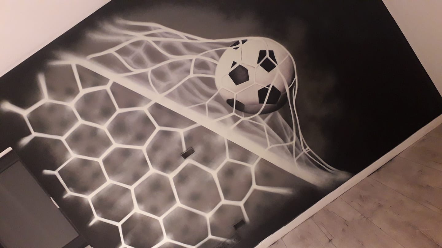 Czarno-biały mural na ścianie przedstawiający piłkę nożną w siatce bramki, wykonany techniką aerografu.