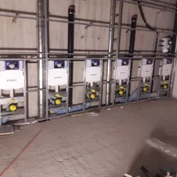 Rząd stelaży podtynkowych Geberit zamontowanych na metalowej konstrukcji w surowym stanie, widoczne rury i elementy instalacji przed zabudową.