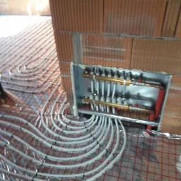 Rozłożone białe rury ogrzewania podłogowego na folii z siatką, widoczny rozdzielacz w metalowej obudowie w ścianie z czerwonej cegły.