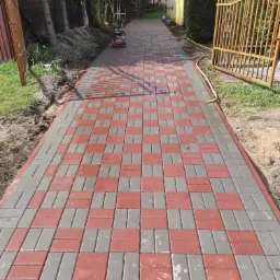 Nowo ułożony chodnik z czerwono-szarej kostki, wzór szachownicy, ubijarka w tle, widoczne świeże obsypanie ziemią po bokach.