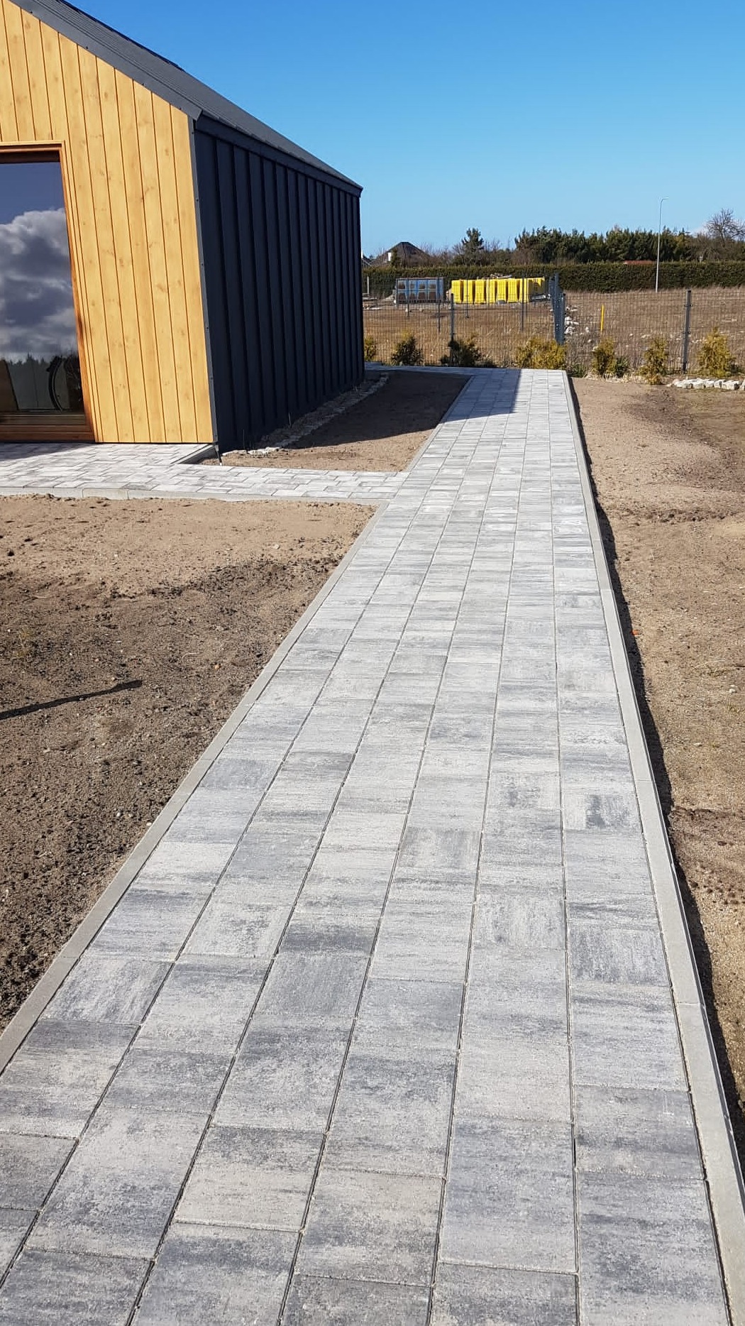 Ścieżka z szarej kostki brukowej prowadząca od nowoczesnego domu z drewnianą elewacją, widok na starannie wyrównany teren i ogrodzenie z siatki.